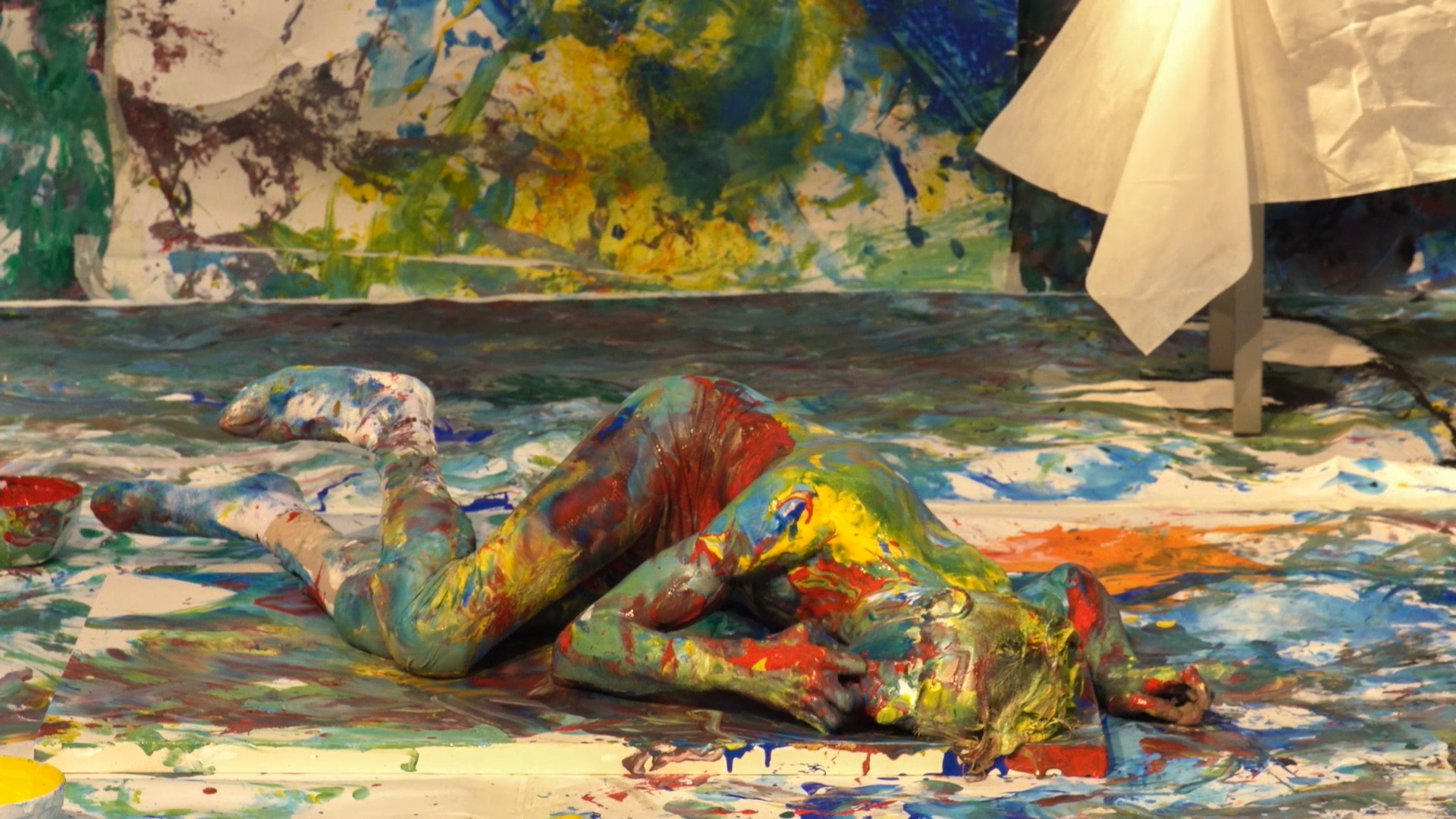 Marcos está deitado de lado em uma tela de pintura. Ele, a tela e o ambiente estão com tinta colorida.